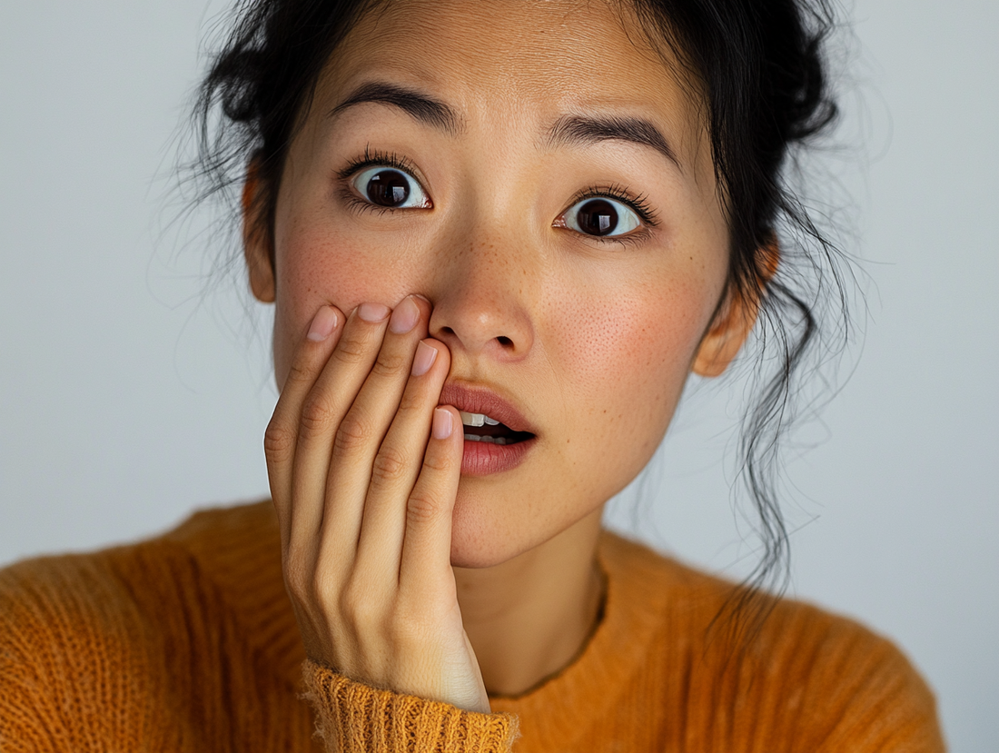 Close-up of an Asian woman holding her hand to the side of her mouth, slightly open and frowning with bad breath. 