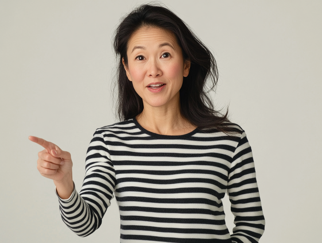 A professional photo of an Asian woman in her late thirties, wearing black and white striped long-sleeved top pointing at something with one finger on plain background.
